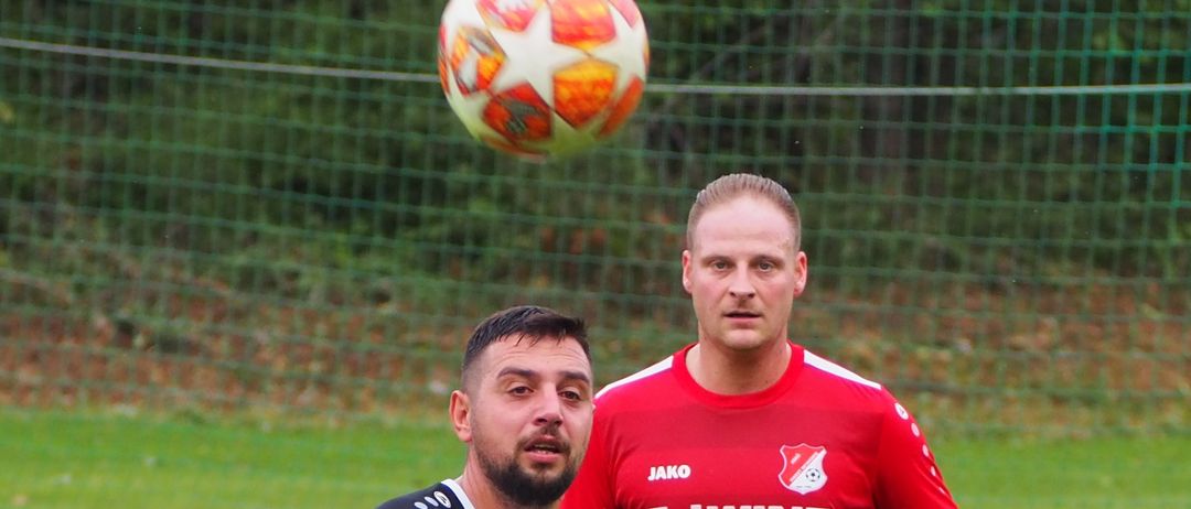 Two soccer players on a field; one wears a white jersey with a sponsor logo, the other a red jersey with a logo and red socks. They are looking in the same direction.