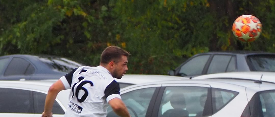 A soccer player in white uniform, with number 16, is jumping to head the ball. Cars are parked in the background. A banner is visible on the side with a logo and some text.