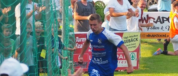 A soccer player in a blue uniform scores a goal while fans cheer and an adult gives high-fives. The net is behind him, and another player is seated nearby.
