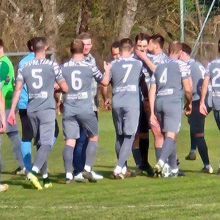 Eine Fußballmannschaft in grauen Trikots mit den Nummern 5, 6, 7, 4 und 2 steht auf einem Feld und diskutiert wahrscheinlich die Strategie.