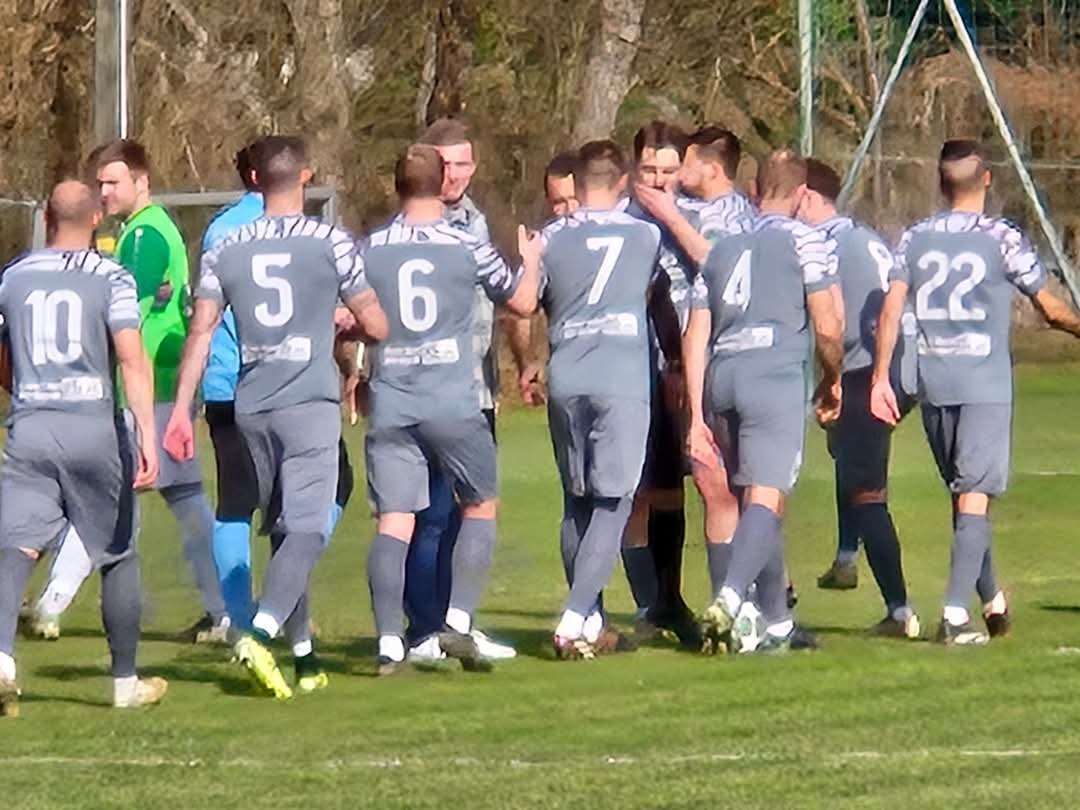 Eine Fußballmannschaft in grauen Trikots mit den Nummern 5, 6, 7, 4 und 2 steht auf einem Feld und diskutiert wahrscheinlich die Strategie.