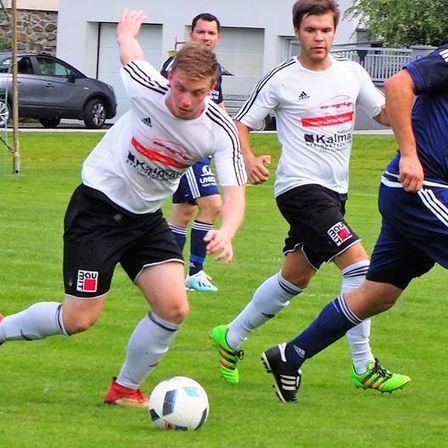 Vier Fußballspieler sind auf einem Feld. Ein Spieler versucht, den Ball zu schießen. Ein anderer Spieler verteidigt. Zwei weitere Spieler sind im Hintergrund.