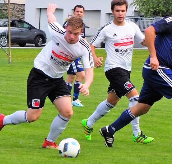 Vier Fußballspieler sind auf einem Feld. Ein Spieler versucht, den Ball zu schießen. Ein anderer Spieler verteidigt. Zwei weitere Spieler sind im Hintergrund.