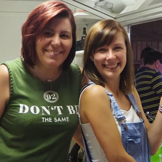 Zwei Frauen lächeln in die Kamera, eine in einem grünen Shirt mit Text und die andere in einer Overall-Jeans. Dahinter steht ein Mann in einem gestreiften Hemd.