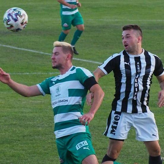 Zwei Fußballspieler konkurrieren auf einem Feld. Ein Spieler trägt grün-weiße Streifen, während der andere schwarze und weiße Streifen trägt. Der Spieler in Grün greift nach dem Ball.