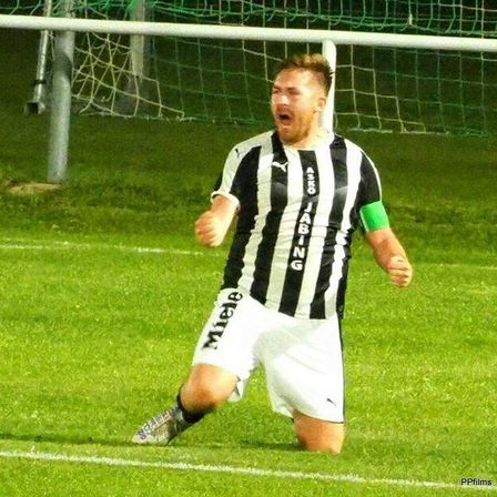 Ein Fußballspieler feiert auf einem Feld mit einem Torpfosten hinter ihm, trägt ein schwarz-weißes Trikot mit 'JABING' und 'Miel' darauf.