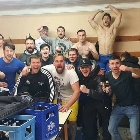 Eine Gruppe von Männern posiert für ein Foto in einem Raum. Einige sind ohne Oberkörper, und ein Mann steht mit erhobenen Armen. Es gibt Kisten mit Bier und einen Tisch im Hintergrund.