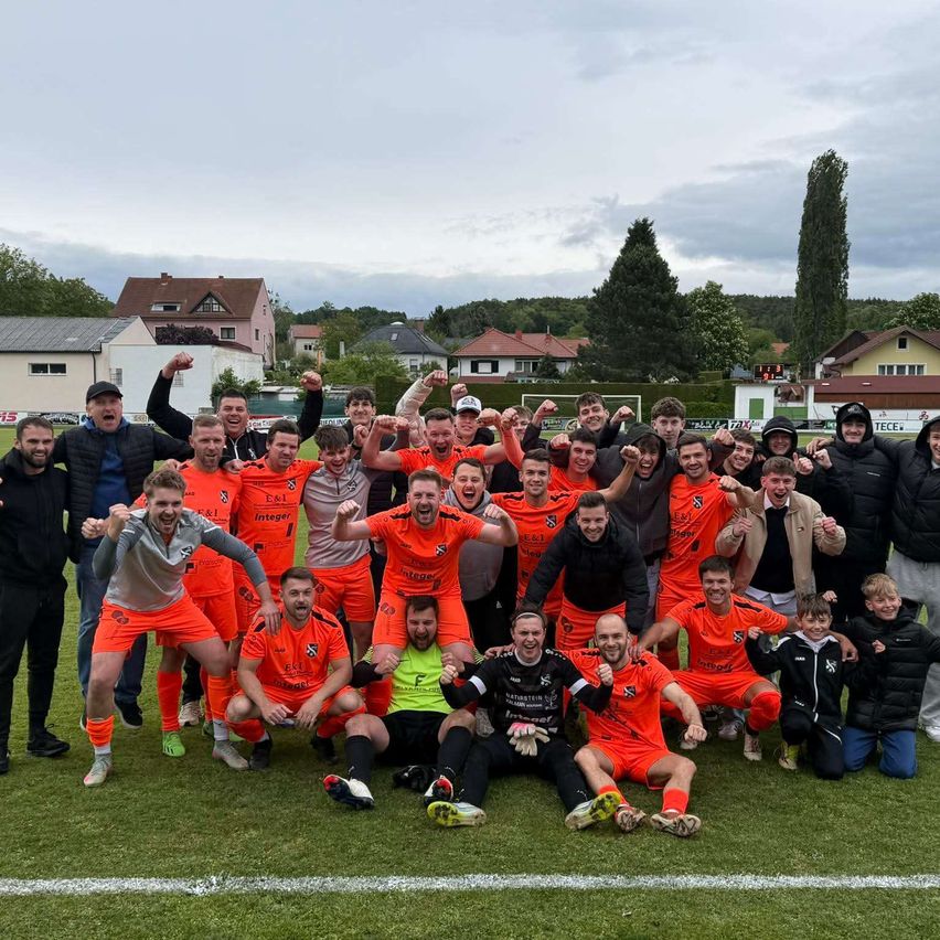 Eine Fußballmannschaft in orangefarbenen Trikots posiert für ein Foto auf einem Feld, mit Fans und einigen mit Mützen im Hintergrund.