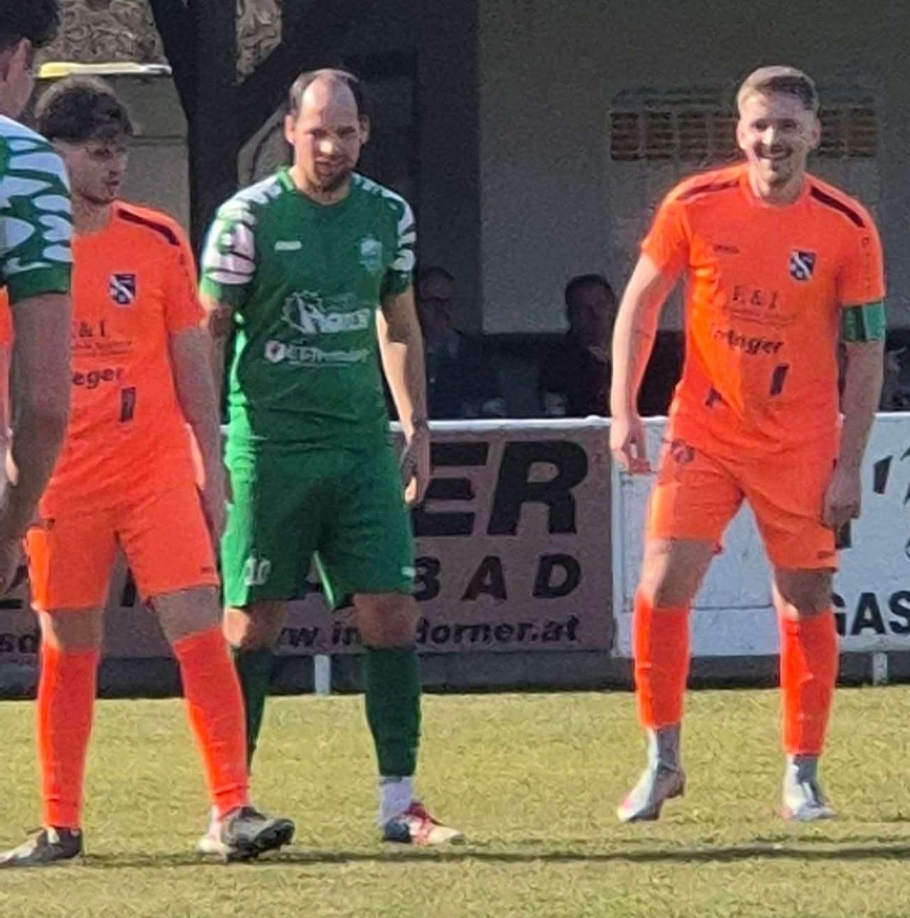 A group of soccer players stands on the field, one wearing a green uniform, another in orange. They appear to be interacting with each other. Behind them, a banner displays text and numbers.