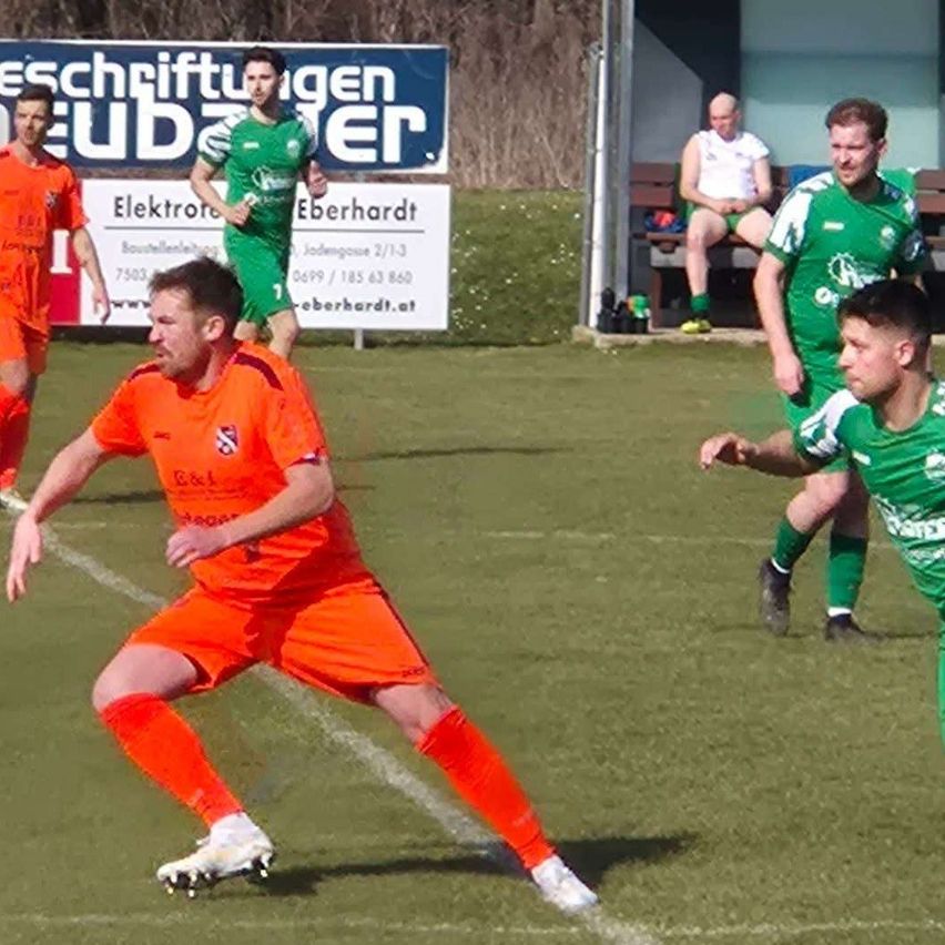 Soccer players in orange and green uniforms play on a field. An advertisement board is in the background.