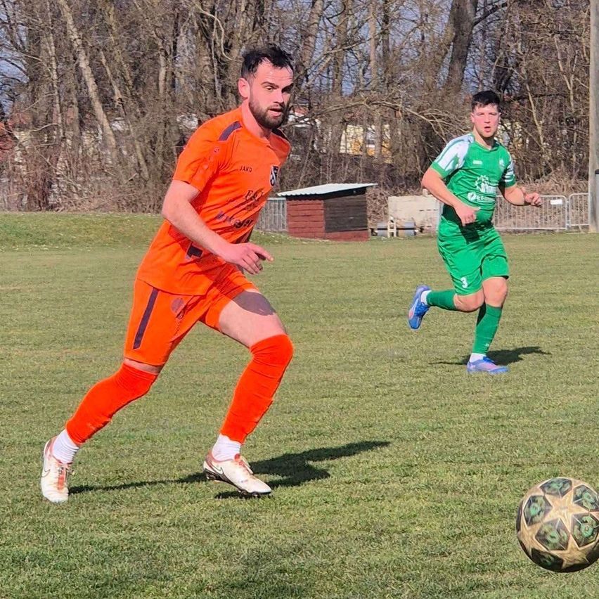 Two soccer players are on a field, one in orange and one in green. The player in orange is running towards the ball. The player in green is chasing the ball. There are trees in the background.