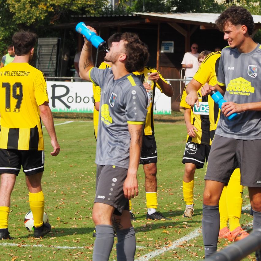 Soccer players in yellow and gray uniforms stand on a field. One player drinks from a blue bottle. A soccer ball is on the ground.