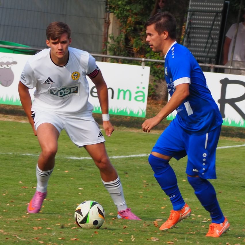 Two soccer players on a field, one in white and the other in blue, are in the midst of a game. The player in white is dribbling the ball while the player in blue is attempting to steal it.