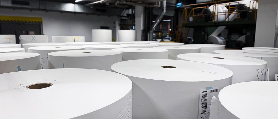 Ein Lagerraum voller zahlreicher großer, weißer Papierrollen, die ordentlich gestapelt sind. Die Rollen haben Barcodes, und das Lager verfügt über Metallträger und Industrieanlagen.