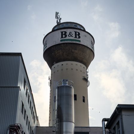 Ein hoher zylindrischer Turm mit einem großen B&B-Logo und Antennen auf der Spitze, umgeben von Industriegebäuden unter einem bewölkten Himmel.