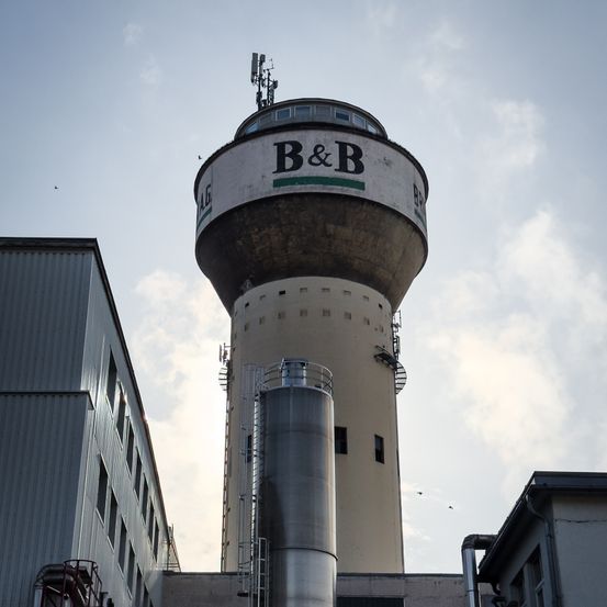 Ein hoher zylindrischer Turm mit einem großen B&B-Logo und Antennen auf der Spitze, umgeben von Industriegebäuden unter einem bewölkten Himmel.