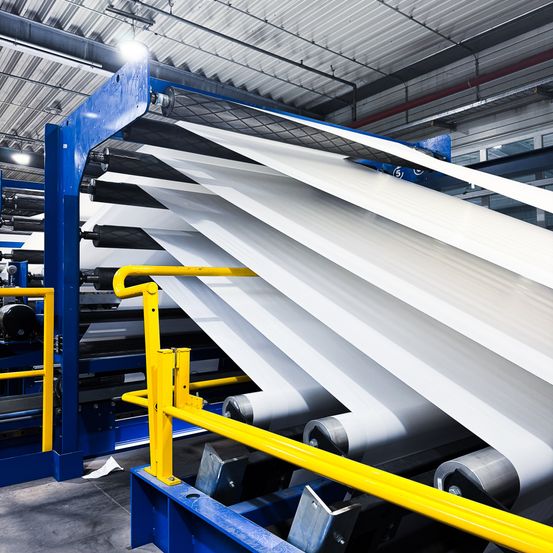 Ein Fabrikinnenraum mit einer blau-gelben Maschine, die große weiße Blätter bearbeitet, unter einer Metalldecke mit Lichtern.