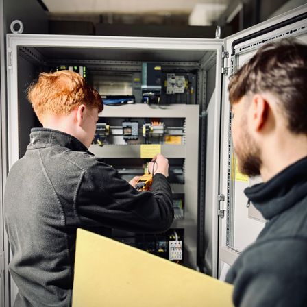 Zwei Männer arbeiten an einem Schaltschrank. Einer hält ein Werkzeug und überprüft die Verkabelung. Der andere beobachtet.