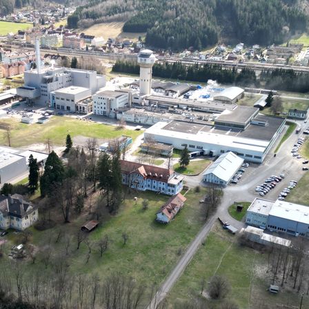 Ein Luftbild eines großen Industriekomplexes mit mehreren Gebäuden, einem hohen Turm und einem dichten Wald. Das Gebiet umfasst Wohnhäuser, einen Fluss und eine Straße mit vielen geparkten Autos.