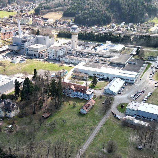 Ein Luftbild eines großen Industriekomplexes mit mehreren Gebäuden, einem hohen Turm und einem dichten Wald. Das Gebiet umfasst Wohnhäuser, einen Fluss und eine Straße mit vielen geparkten Autos.