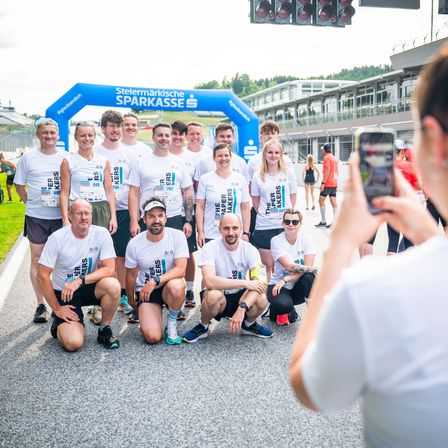 Eine Gruppe von Menschen in weißen T-Shirts posiert für ein Foto an der Startlinie eines Rennens. Die Frau rechts macht ein Foto mit ihrem Handy.