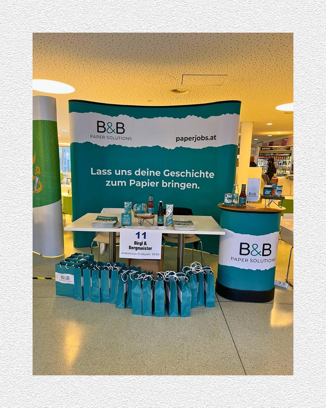 Ein Stand von B&B Paper Solutions mit Werbetüten und einem Tisch mit Flaschen und Büchern. Ein Banner lautet: 'Lass uns deine Geschichten zum Papier bringen.'