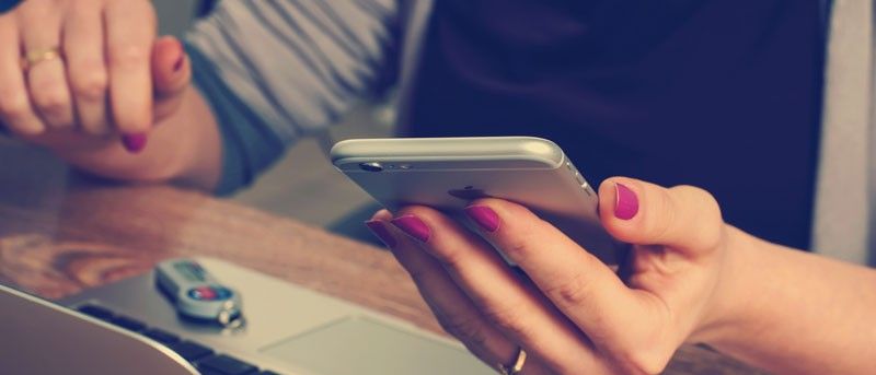 Eine Person mit lackierten Nägeln benutzt ein Handy neben einem Laptop auf einem Schreibtisch.