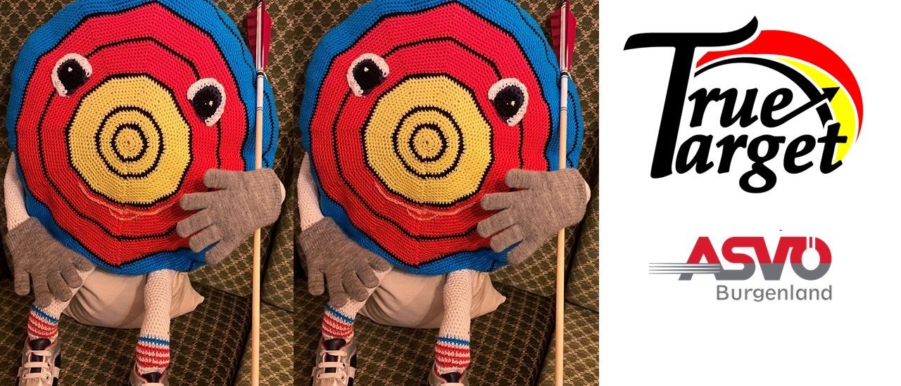 Eine Puppe mit einem gehäkelten Zieldesign, die zwei Pfeile hält, sitzt auf einer Couch. Die Puppe trägt graue Handschuhe, gestreifte Socken und Turnschuhe.