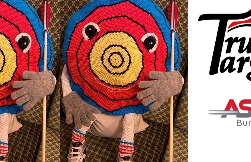 Eine Puppe mit einem gehäkelten Zieldesign, die zwei Pfeile hält, sitzt auf einer Couch. Die Puppe trägt graue Handschuhe, gestreifte Socken und Turnschuhe.