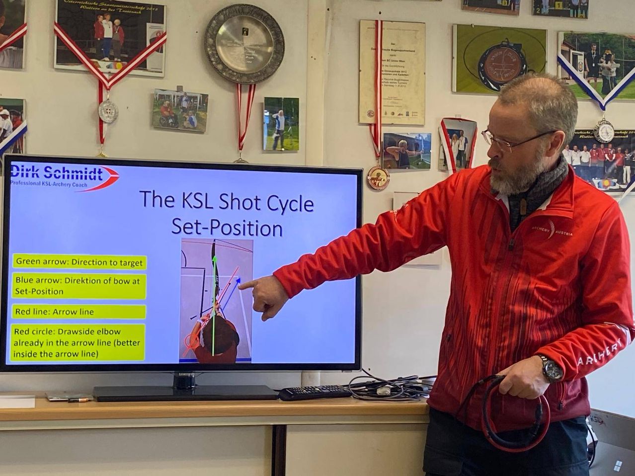 Ein Mann zeigt mit dem Finger auf einen Monitor, der 'The KSL Shot Cycle Set-Position' anzeigt. Der Bildschirm zeigt eine Illustration von Bogen und Pfeilen. Er trägt einen roten Mantel und eine Brille. Die Wand hinter ihm ist mit Medaillen und gerahmten Bildern dekoriert.