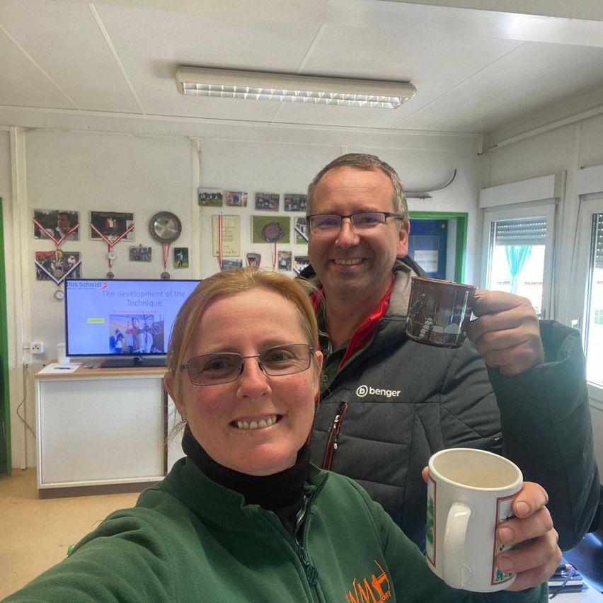 Ein Mann und eine Frau lächelnd, halten Tassen in der Hand, stehen vor einem Monitor, der eine Präsentation über die Entwicklung der Technik zeigt. Dahinter ein Raum mit verschiedenen Dekorationen und einer Uhr an der Wand.