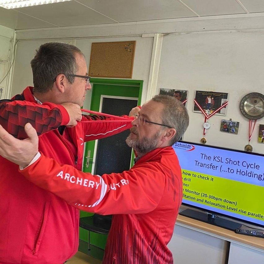 Zwei Männer in roten Jacken üben Bogenschießen drinnen. Einer hält den Arm des anderen, während er zielt. Ein Monitor zeigt den KSL Shot Cycle Transfer.