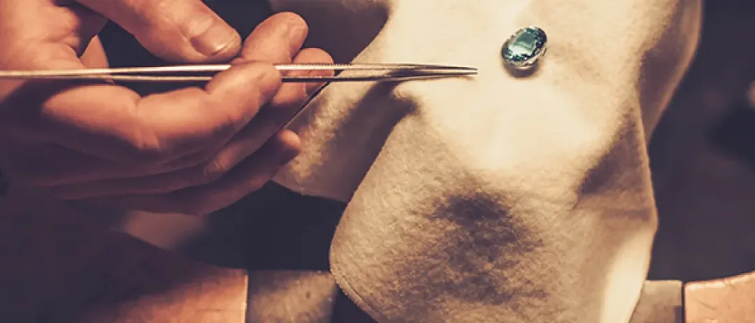 Ein Juwelier hält eine Pinzette mit einem blauen Edelstein auf einem Tuch. Eine Lupe und andere Werkzeuge liegen in der Nähe.