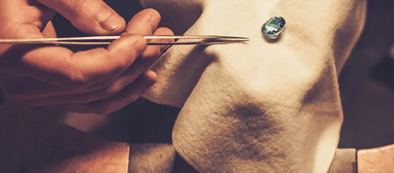 Ein Juwelier hält eine Pinzette mit einem blauen Edelstein auf einem Tuch. Eine Lupe und andere Werkzeuge liegen in der Nähe.