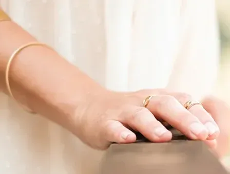 Eine Person in einem weißen Kleid hält mit ihren Händen ein Holzgeländer fest, geschmückt mit goldenen Ringen und Armbändern.