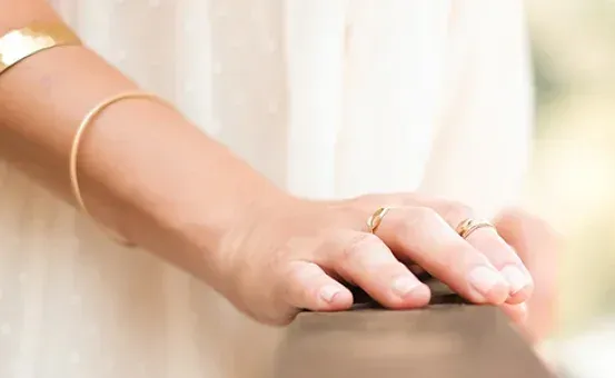 Eine Person in einem weißen Kleid hält mit ihren Händen ein Holzgeländer fest, geschmückt mit goldenen Ringen und Armbändern.