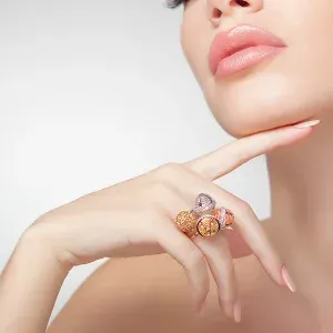 Eine Frau mit rosa Lippen und gepflegten Nägeln zeigt auf einen Ring an ihrem Finger. Der Ring hat einen farbenfrohen Edelstein.