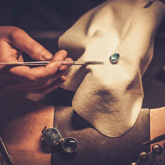 Eine Person verwendet eine Pinzette, um einen blauen Edelstein auf ein Tuch zu legen. Eine Lupe und Werkzeuge sind in der Nähe.