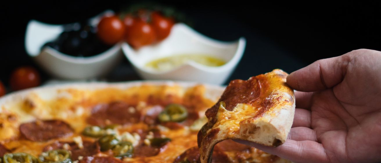 Eine Person hält ein Stück Pizza mit Peperoni und Jalapenos. Im Hintergrund befinden sich Schalen mit Oliven und Tomaten.