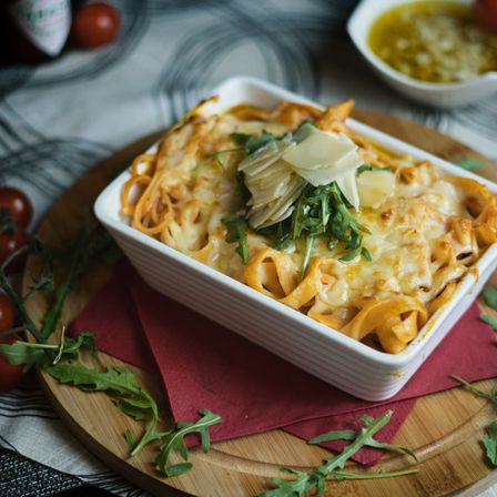 Ein quadratisches weißes Gericht mit Käse überzogener Pasta steht auf einem Holzteller, garniert mit grünen Kräutern und Käse. Umgeben ist es von Tomaten, Rucola und einer kleinen Schüssel Sauce.