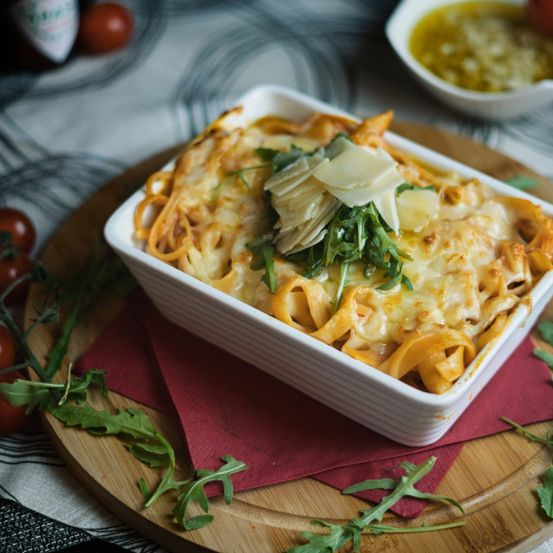 Ein quadratisches weißes Gericht mit Käse überzogener Pasta steht auf einem Holzteller, garniert mit grünen Kräutern und Käse. Umgeben ist es von Tomaten, Rucola und einer kleinen Schüssel Sauce.