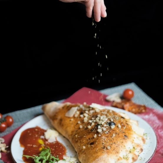 Eine Hand streut Käse über ein Calzone mit Tomatensoße auf einem Teller, auf einem roten Tuch platziert.