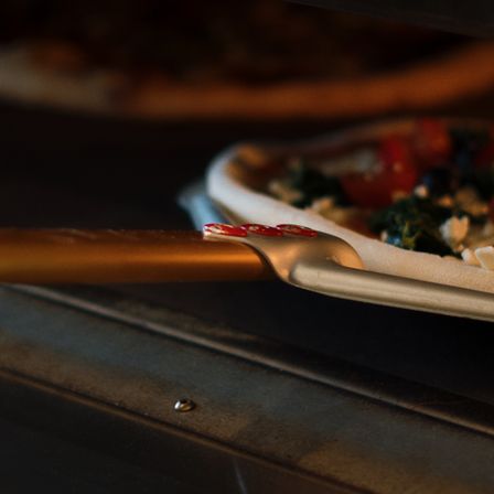 Eine Pizza mit Tomaten, Spinat und Käse wird aus einem Ofen mit einem goldenen Pizzaschaufel entfernt.