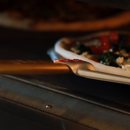 Eine Pizza mit Tomaten, Spinat und Käse wird aus einem Ofen mit einem goldenen Pizzaschaufel entfernt.