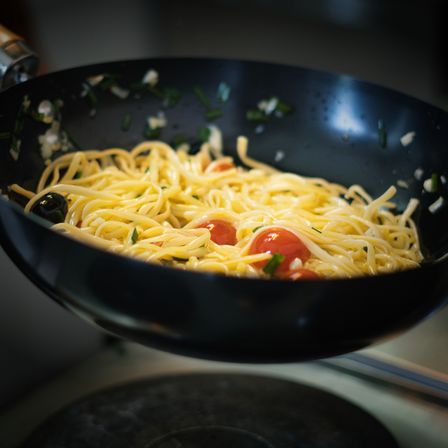 Ein schwarzer Wok gefüllt mit Pasta, garniert mit geschnittenen Tomaten und Oliven, wird von einem Griff angehoben und steht auf einem Herd.