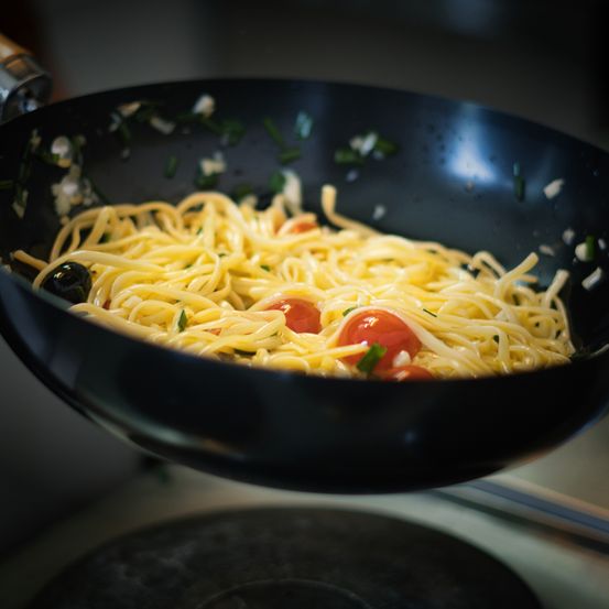 Ein schwarzer Wok gefüllt mit Pasta, garniert mit geschnittenen Tomaten und Oliven, wird von einem Griff angehoben und steht auf einem Herd.