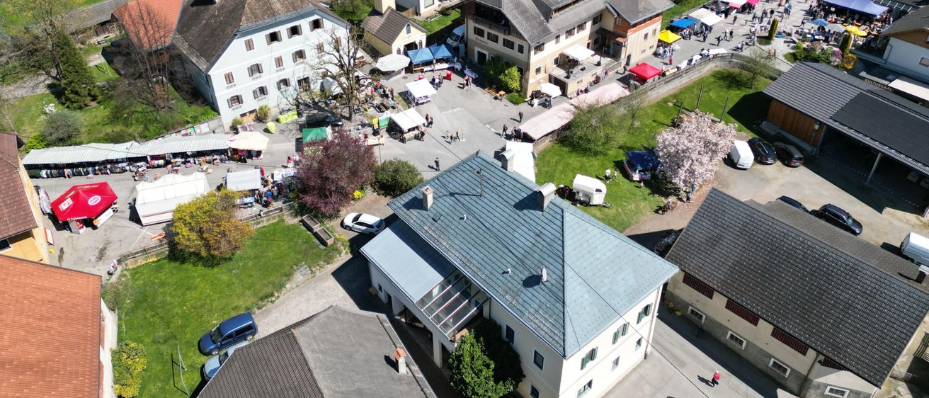 Luftaufnahme eines kleinen Dorfes mit verschiedenen Häusern, einer Kirche und einem Markt mit vielen Zelten und Menschen.
