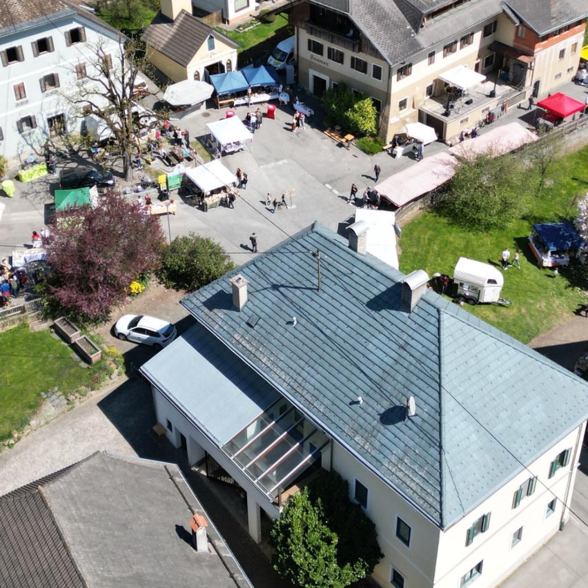 Luftaufnahme eines kleinen Dorfes mit verschiedenen Häusern, einer Kirche und einem Markt mit vielen Zelten und Menschen.