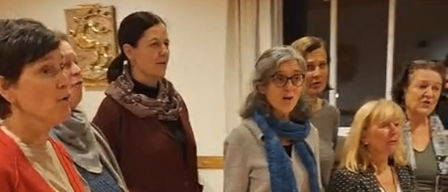 Eine Gruppe von Frauen, alle in Winterkleidung gekleidet, stehen in einem Raum und scheinen zu singen. Sie stehen auf einem Holzboden.