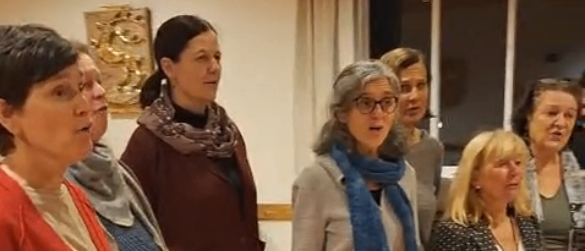 Eine Gruppe von Frauen, alle in Winterkleidung gekleidet, stehen in einem Raum und scheinen zu singen. Sie stehen auf einem Holzboden.